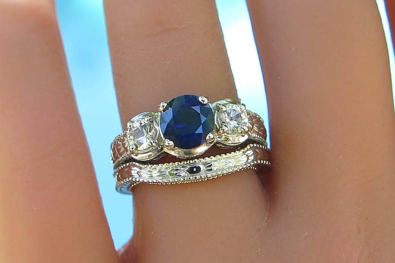 A close-up of a hand wearing a three-stone ring set. The center ring features an oval-cut deep blue sapphire flanked by two round clear white sapphires, set in a white gold band with engraving.