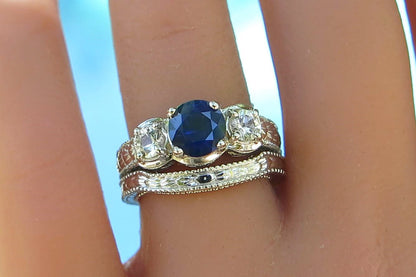 A close-up of a hand wearing a three-stone ring set. The center ring features an oval-cut deep blue sapphire flanked by two round clear white sapphires, set in a white gold band with engraving.