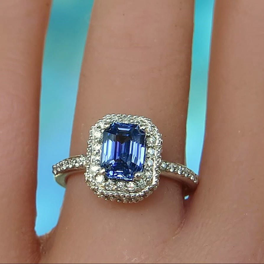 A white gold ring with a rectangular emerald-cut blue sapphire center stone and a halo of small clear stones, shown worn on a finger against a bright blue background.