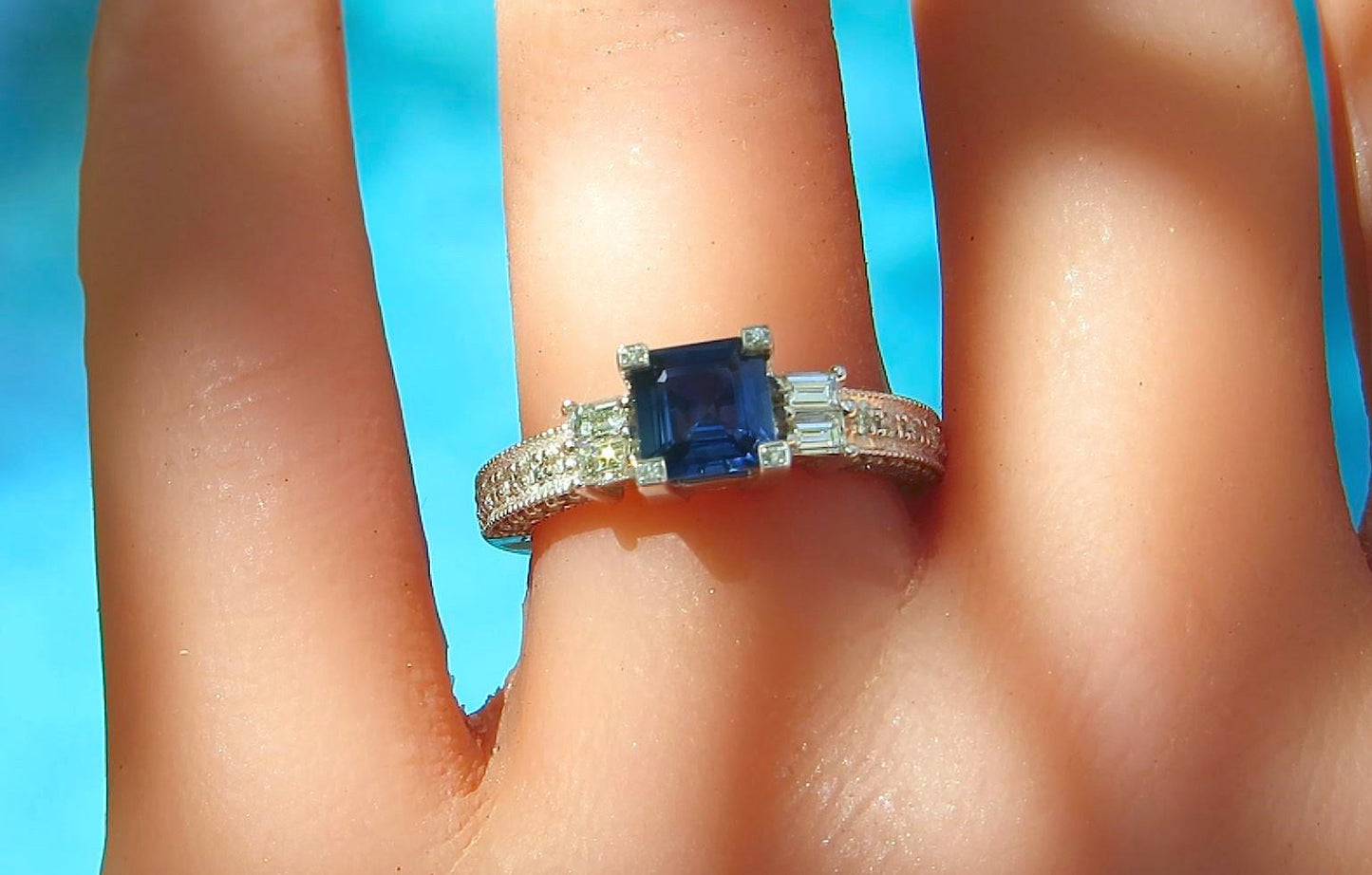 A close-up of a ring worn on a finger, featuring a square-cut deep blue center stone with baguette and round side stones on a silver-toned band, set against a bright blue background.