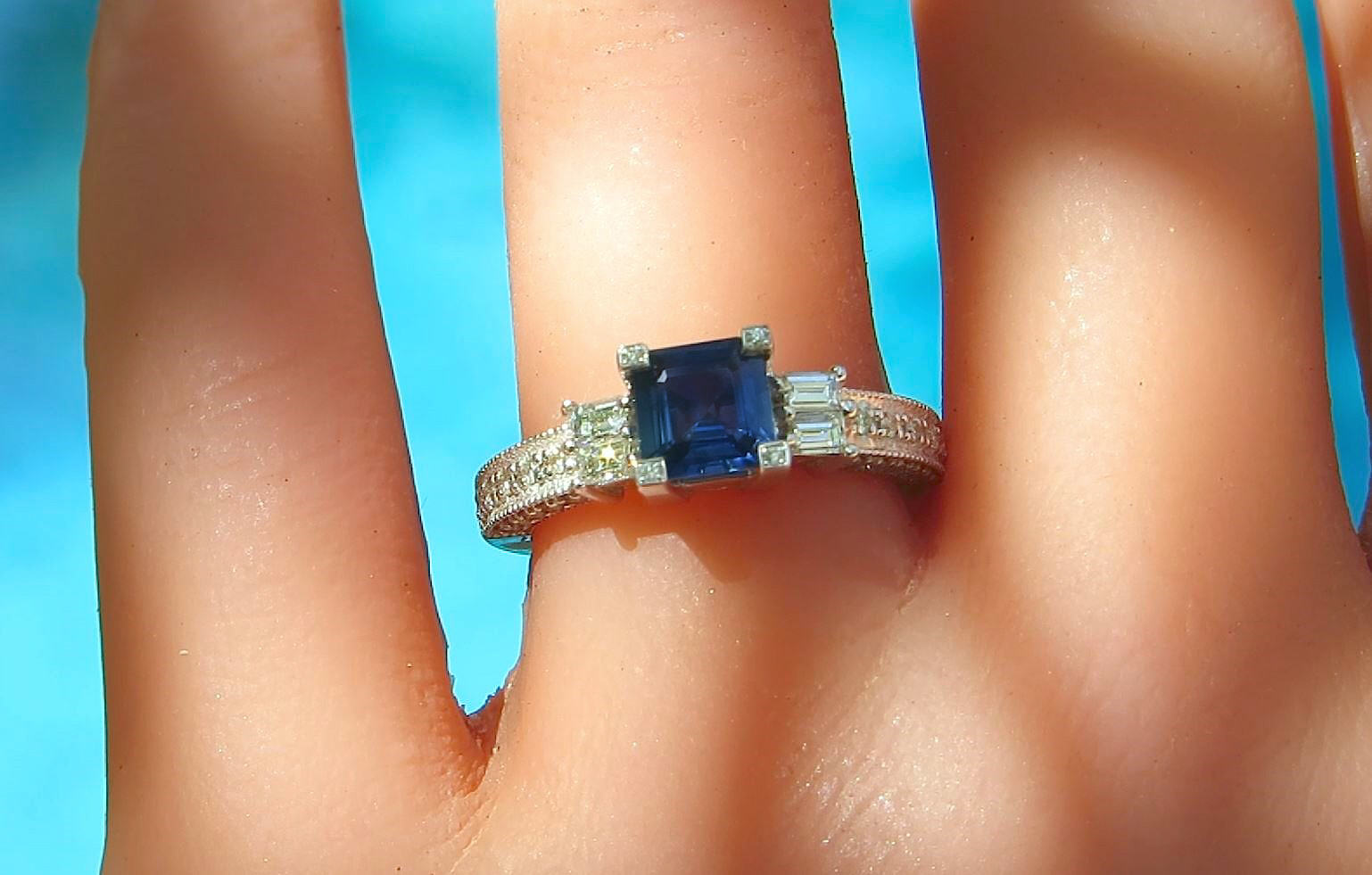 A close-up of a ring worn on a finger, featuring a square-cut deep blue center stone with baguette and round side stones on a silver-toned band, set against a bright blue background.