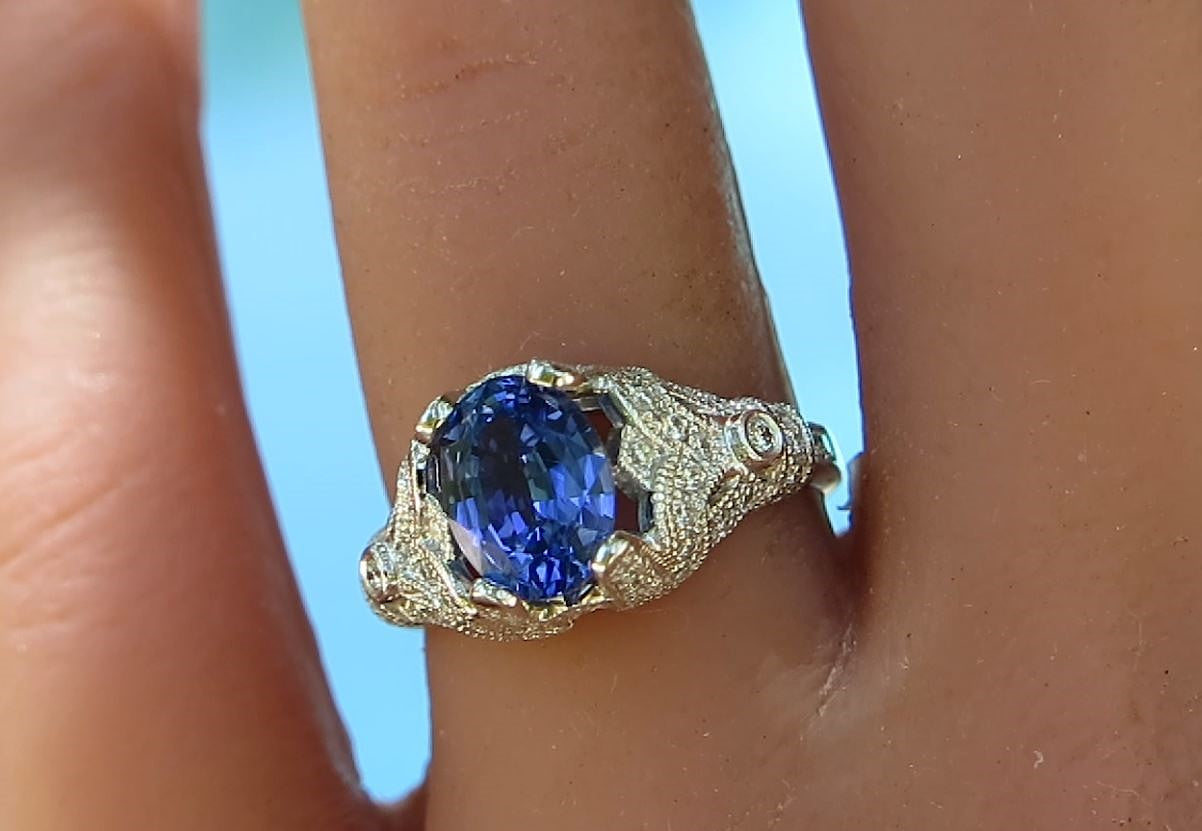 A close-up of a ring worn on a finger, featuring an oval deep blue gemstone in an ornate white gold setting with small clear accent stones.