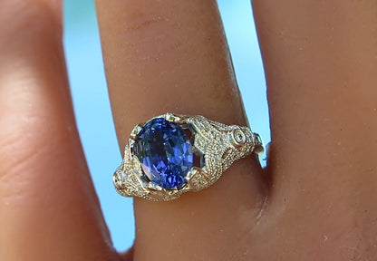 A close-up of a ring worn on a finger, featuring an oval deep blue gemstone in an ornate white gold setting with small clear accent stones.