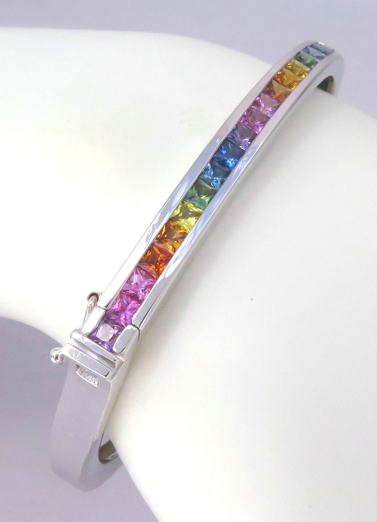 A white gold cuff bracelet featuring a channel set row of rainbow-colored sapphires, displayed on a white curved jewelry stand.