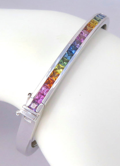 A white gold cuff bracelet featuring a channel set row of rainbow-colored sapphires, displayed on a white curved jewelry stand.