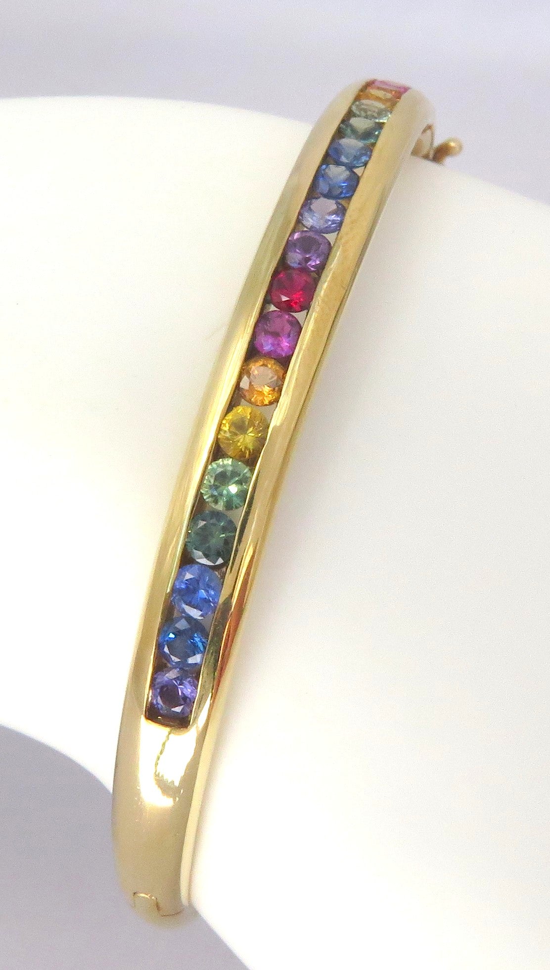 A gold bangle bracelet featuring a channel of round, rainbow-colored sapphires, displayed on a white mannequin arm.