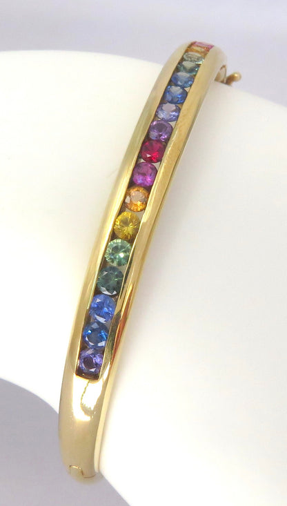A gold bangle bracelet featuring a channel of round, rainbow-colored sapphires, displayed on a white mannequin arm.